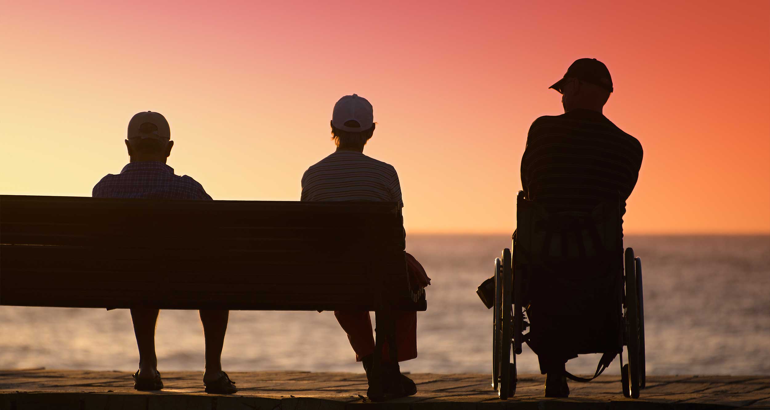 Two people sitting on a bench and one wheelchair user watching the sunset.