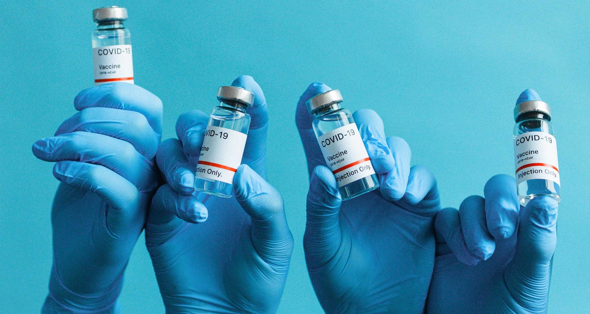 Vials of vaccines, held by hands covered with blue gloves.