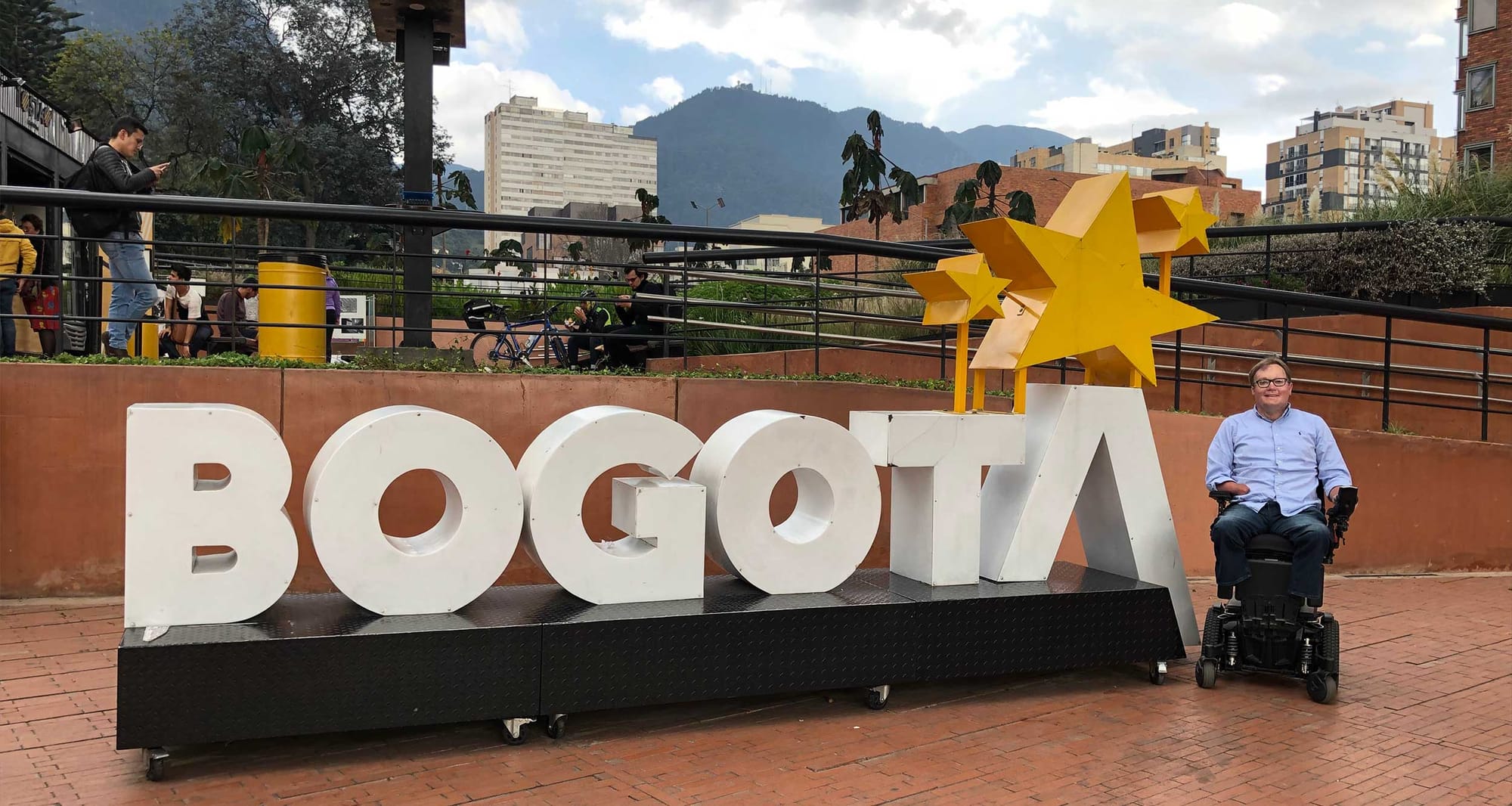 John seated in wheelchair next to Bogota sign, made with large white letters.