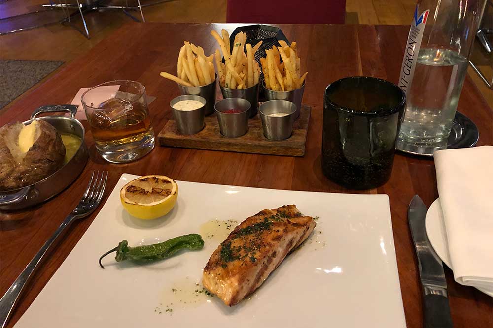 Salmon, baked potato, french fries, glass of bourbon and water bottle on table.