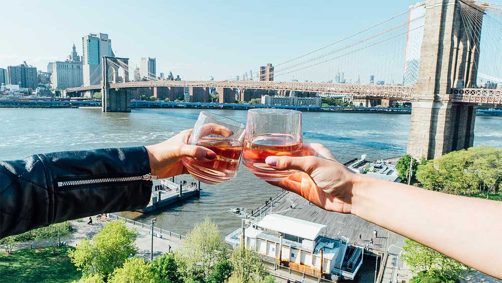 Two people clinking glasses from rooftop in front of NYC skyline.