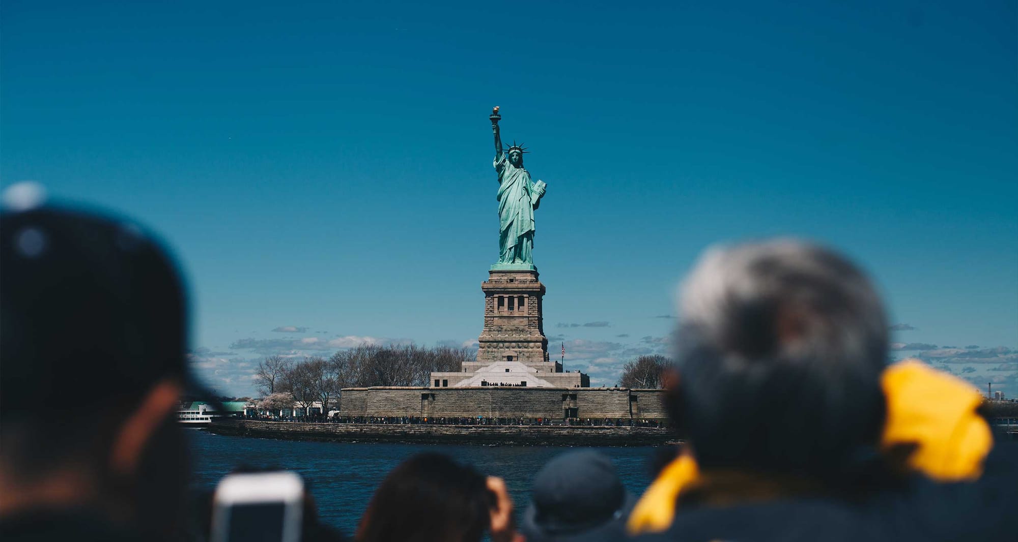 Crowd of people looking at and taking photos of the Statue of Liberty, seen on an island in the distance.