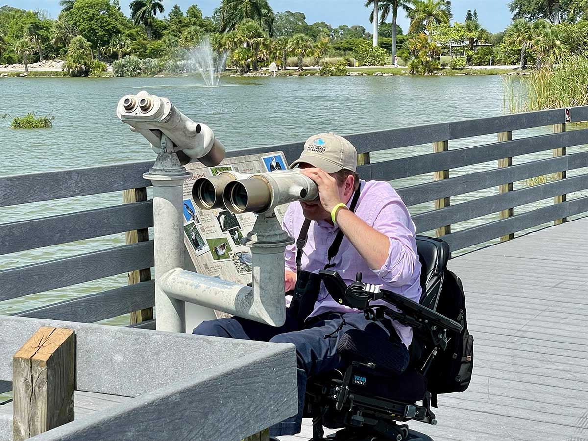 John in wheelchair looking through viewfinder at pier over lake.