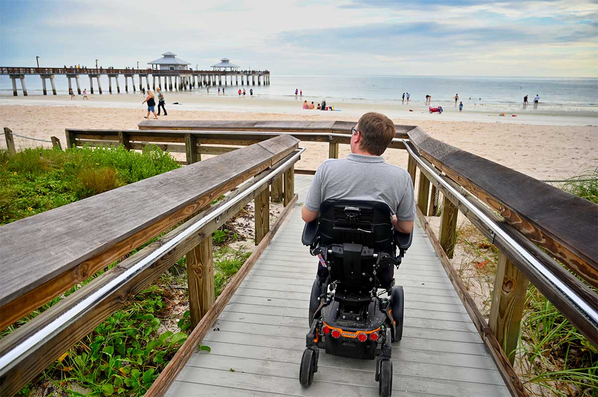 John rolling in wheelchair on boardwalk towards beach with tall pier in distance.