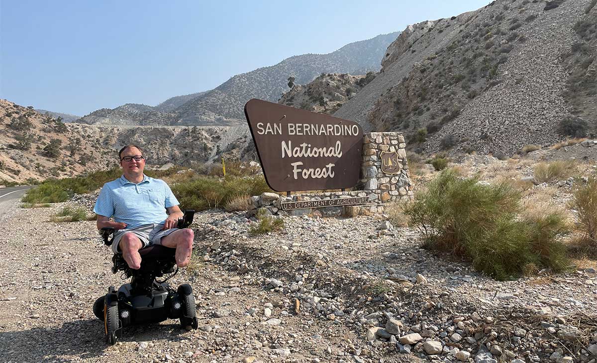 John sitting in wheelchair in front of San Bernardino National Forest sign.