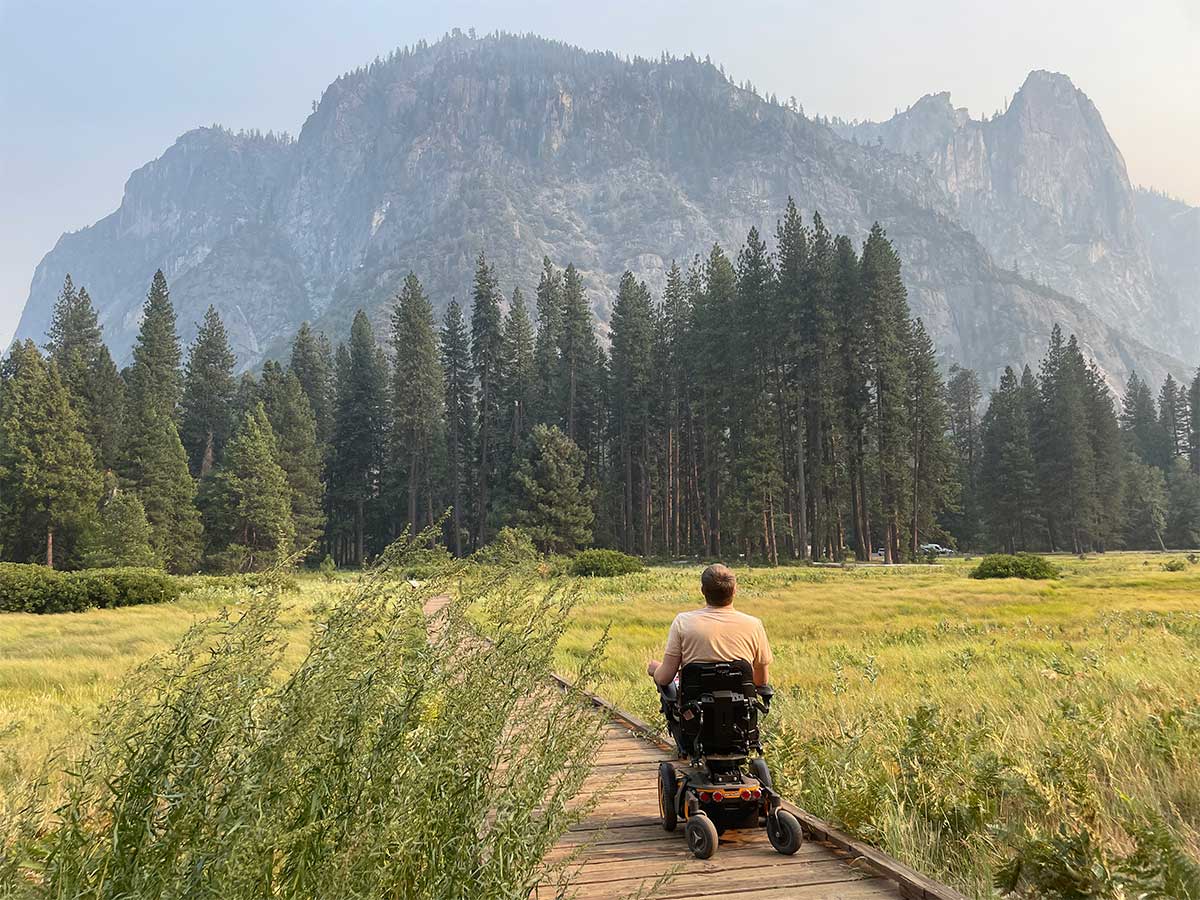 John in wheelchair rolling on wooden boardwalk trail through field.