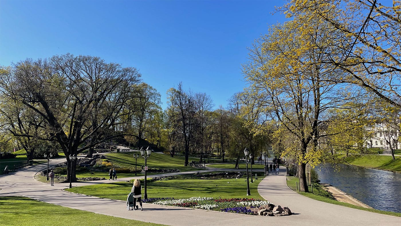 Paved pathways leading in multiple directions through a lush green park with a water body and fountain in the center.