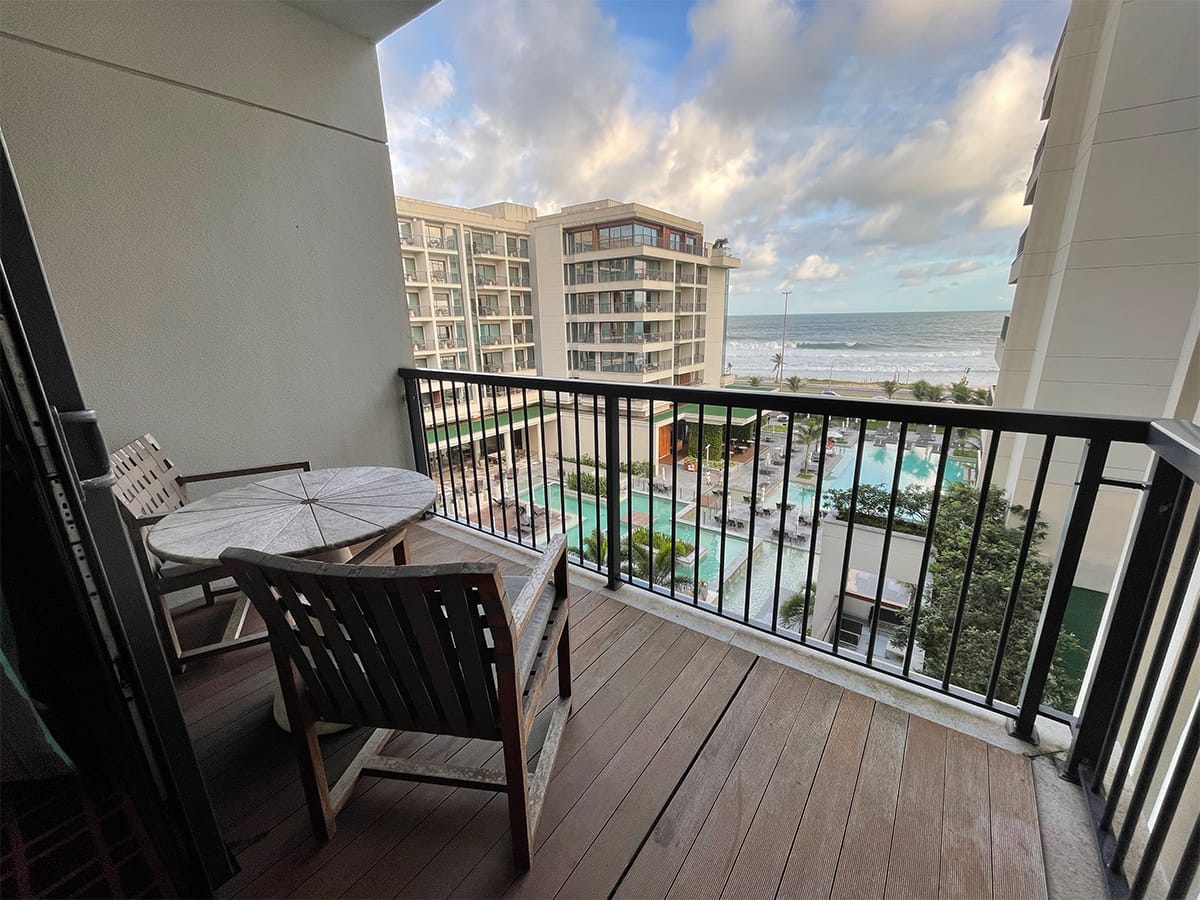 Balcony overlooking the swimming pool and beach, with two chairs and a table.