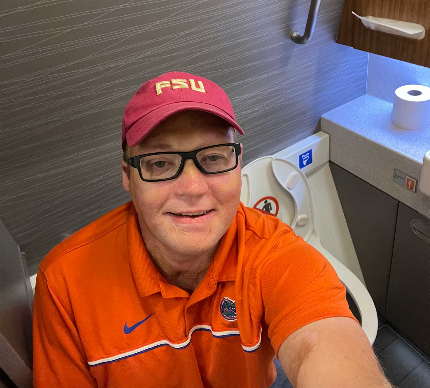 John seated on the aisle chair inside of the accessible lavatory.