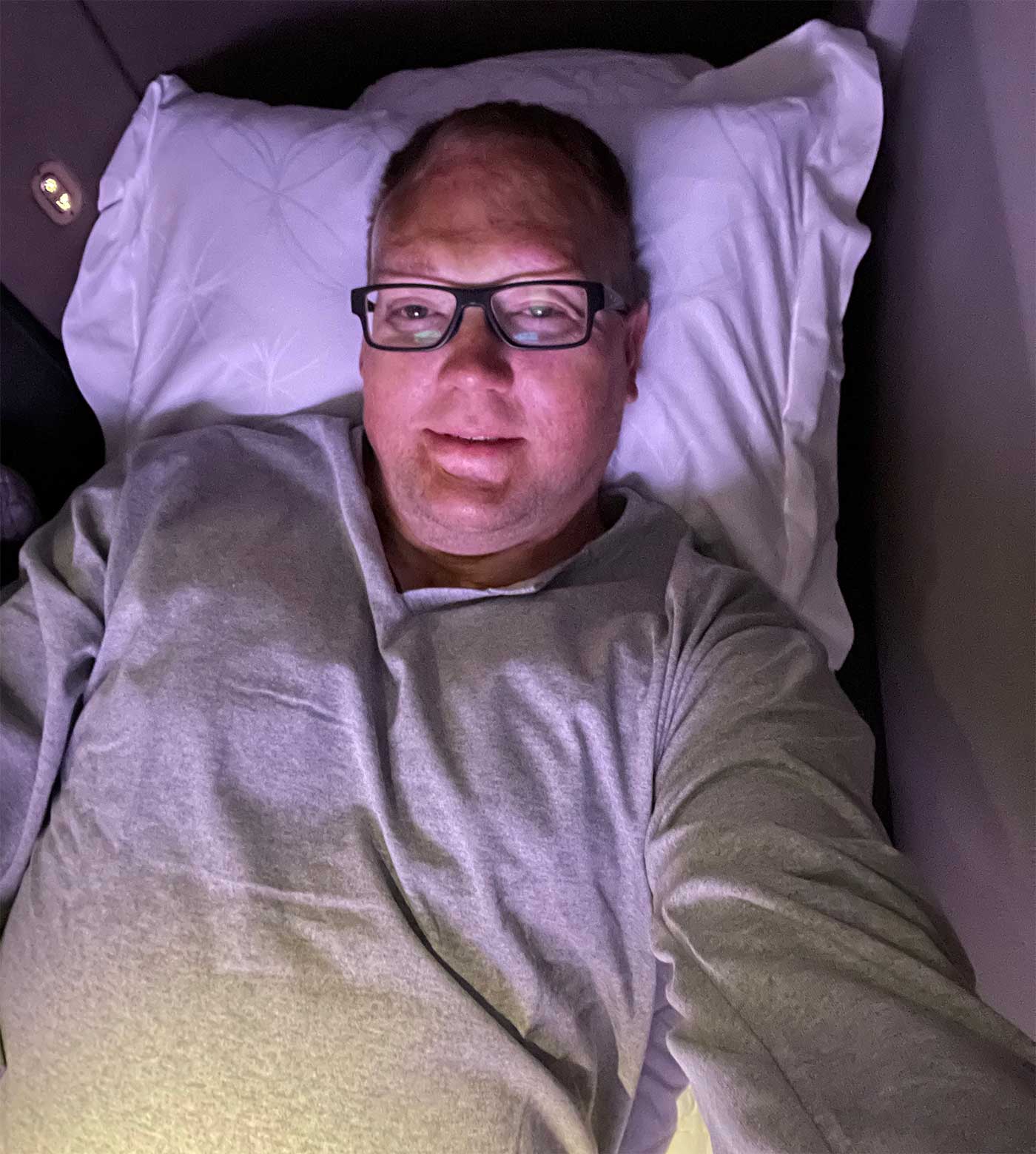 Selfie of John laying in bed with a fluffy pillow and Qatar Airways pajamas.