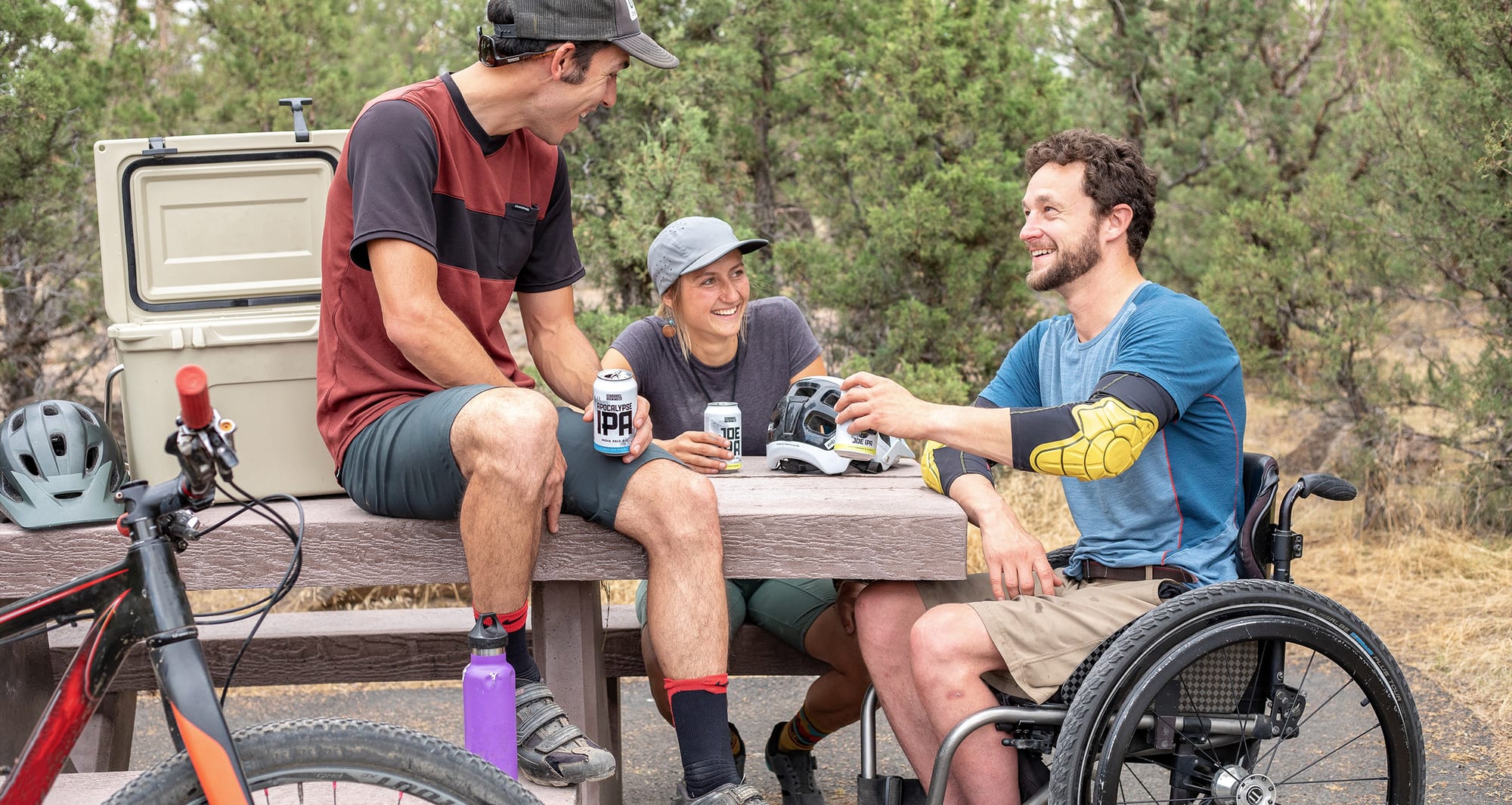 Wheelchair user sitting outdoors with two friends at a picnic table in a park.