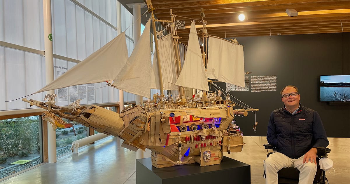 John in his wheelchair next to a sailboat made with cardboard.