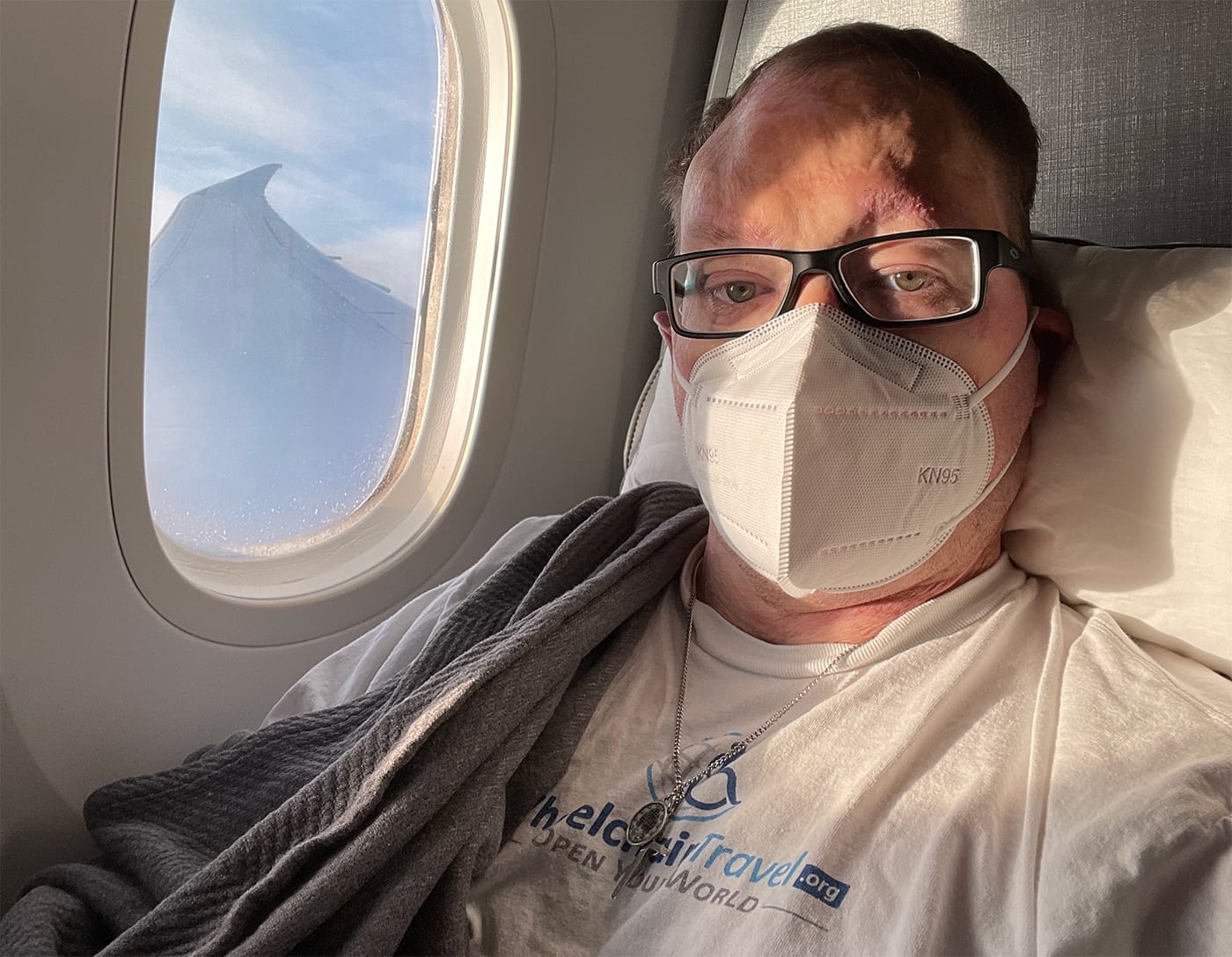 John seated in an airplane seat during flight while wearing a Wheelchair Travel logo t-shirt.