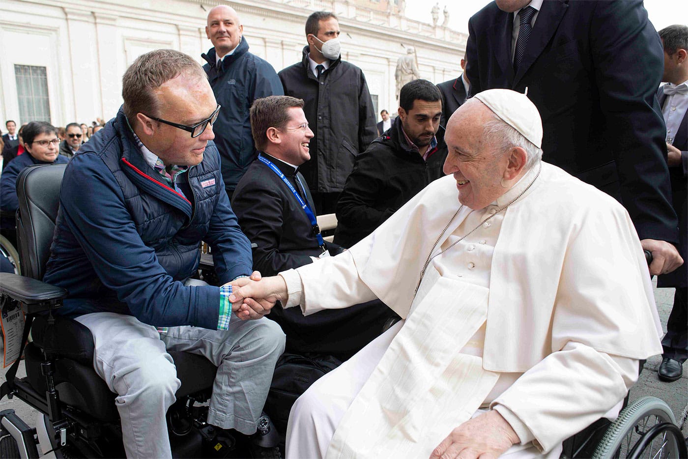 John seated in his power wheelchair shaking hands with Pope Francis, who is seated in a manual wheelchair.