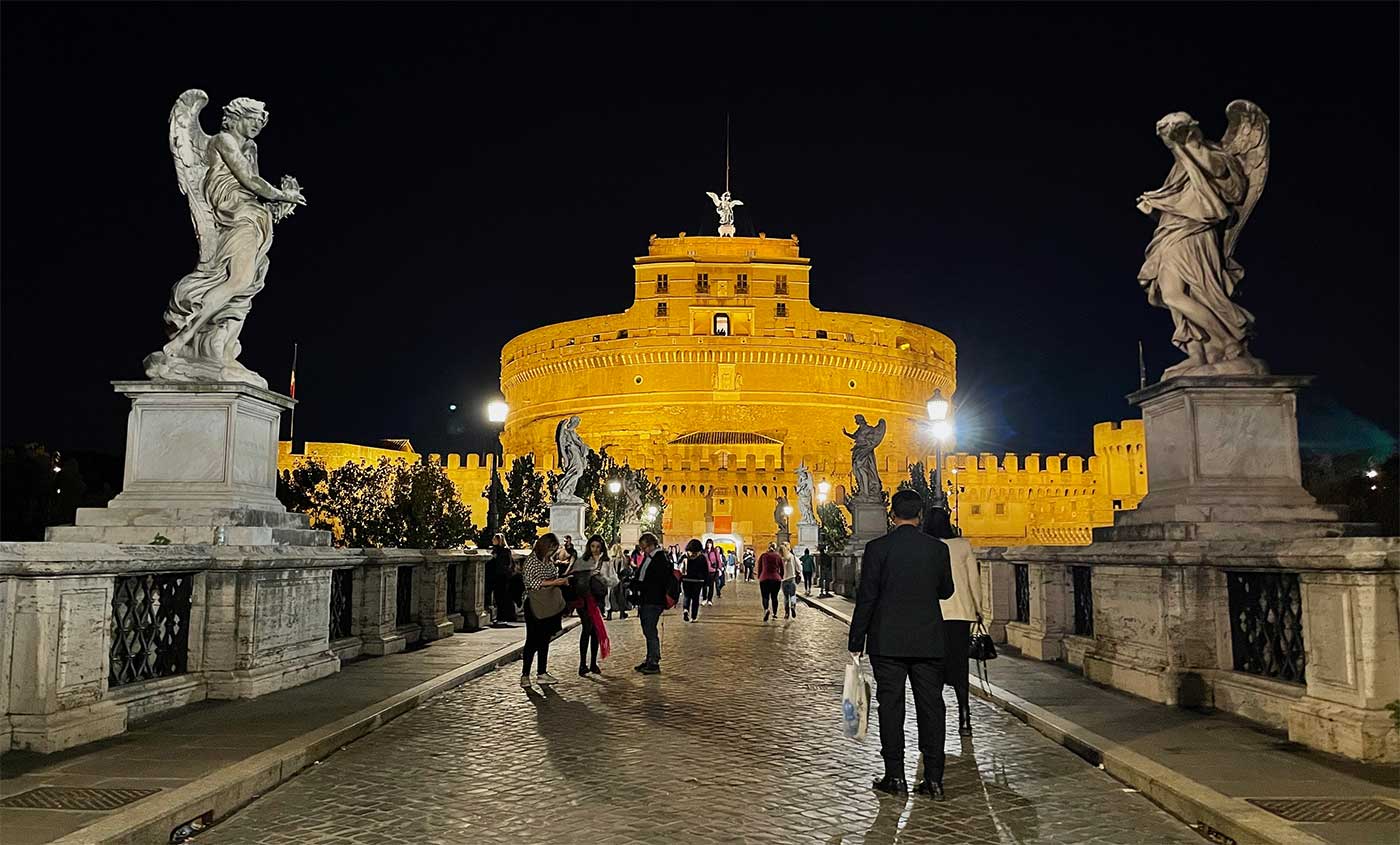 Round castle lit up at night, seen from a bridge with two large angel statues on each side of the bridge.