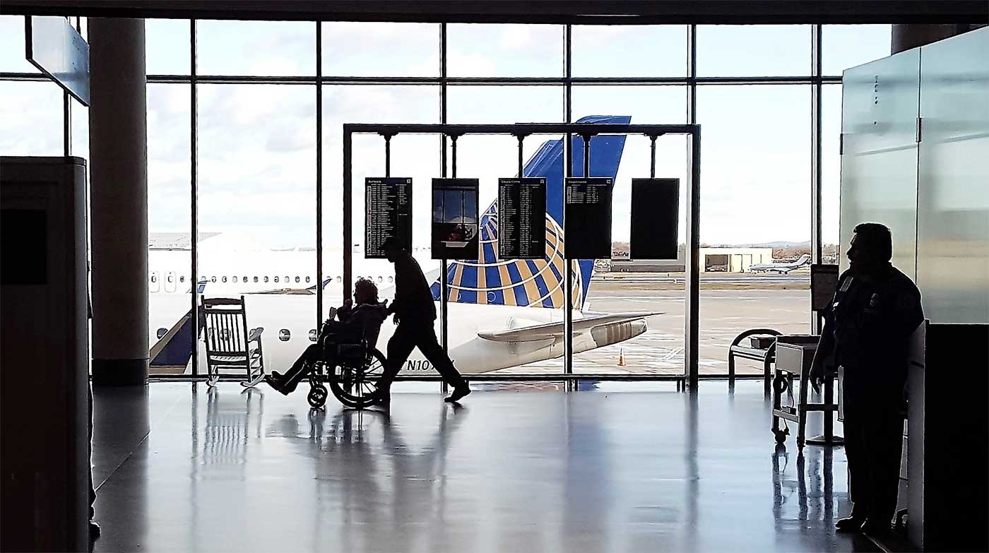 Person pushing a wheelchair user in front of an airport window with a United Airlines jet outside.