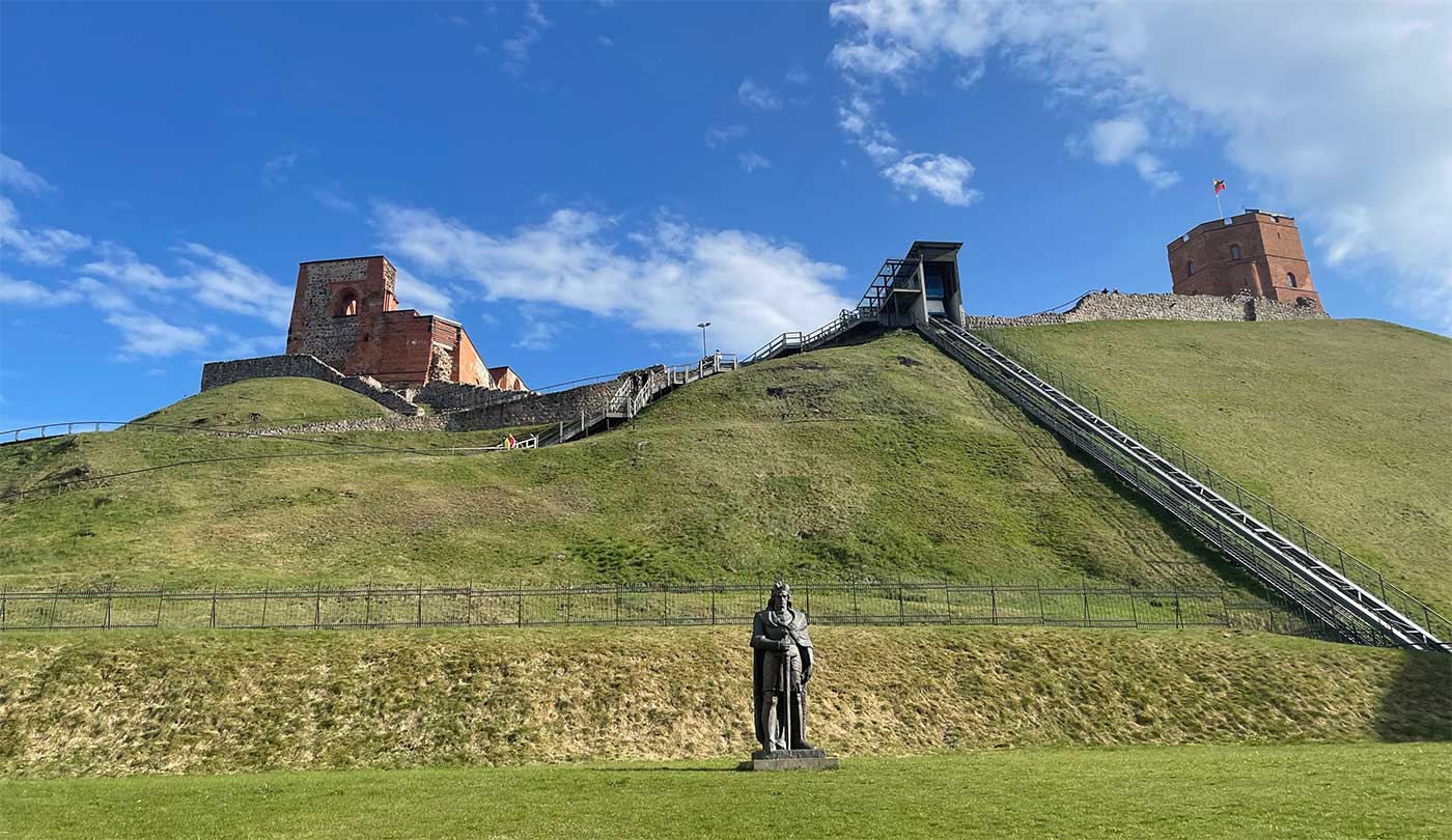 Brick stone castle tower on top of tall grassy hill, with funicular tracks going up the hillside and a statue of a king at its base.