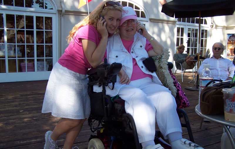 Woman standing next to and embracing her mother, who is seated in a power wheelchair.