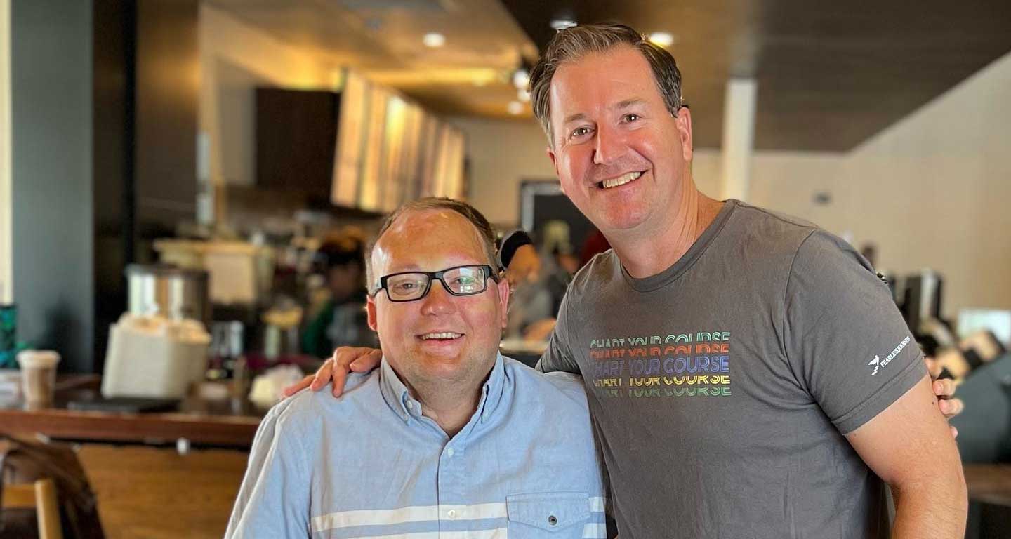 John seated in his wheelchair next to his friend at a Starbucks.