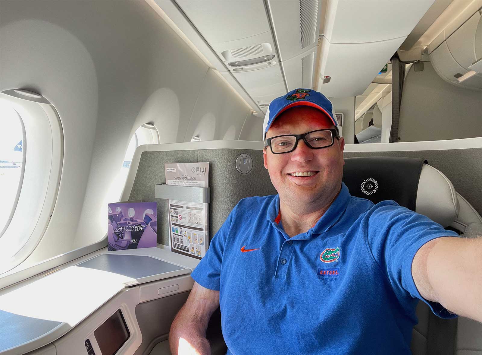 Selfie of John wearing blue polo shirt and hat seated in business class on a Fiji Airways airplane.