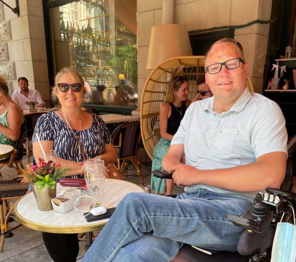 John seated in his wheelchair at a table with his friend.