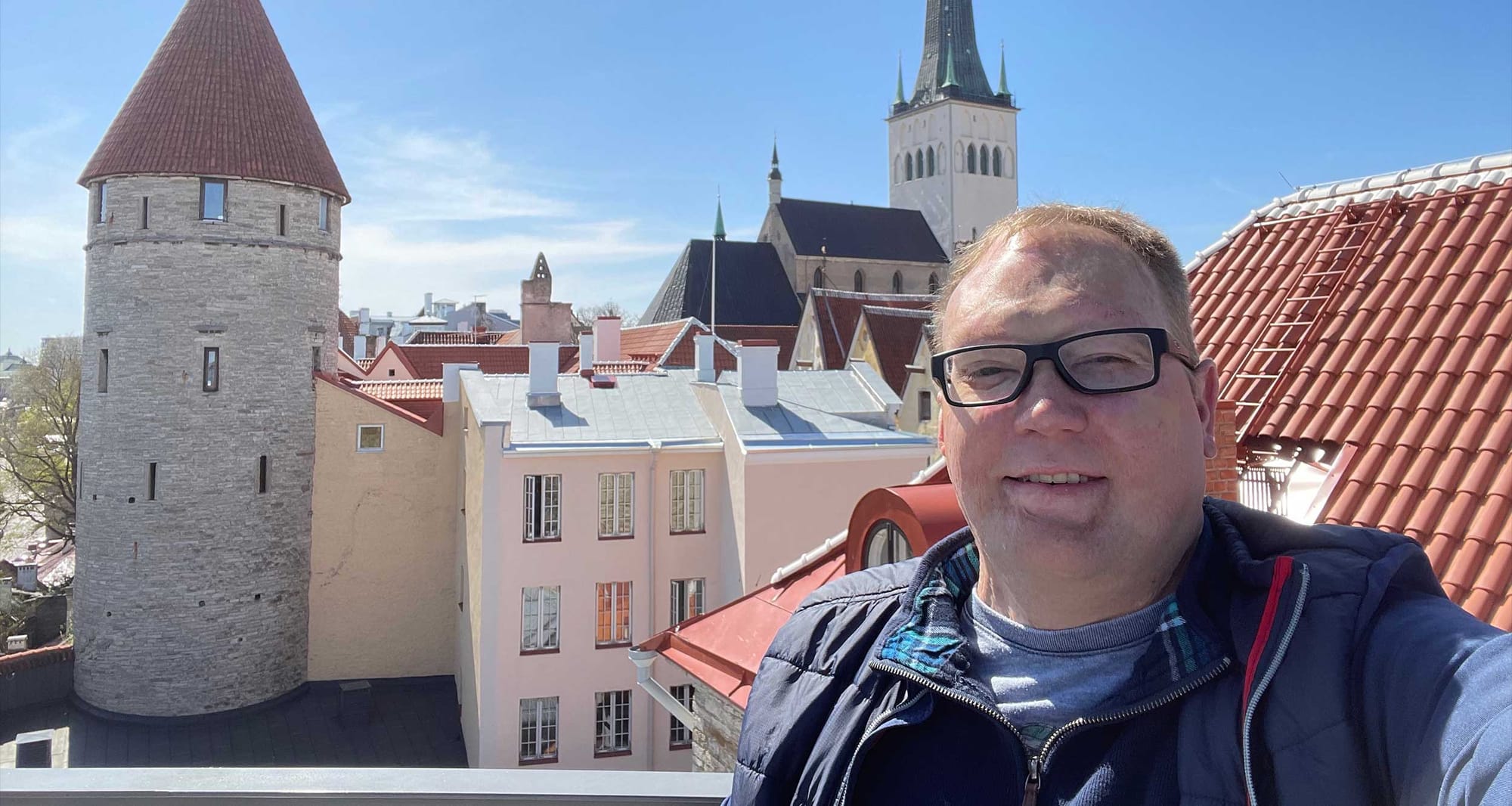 Selfie of wheelchair user on rooftop in front of Tallinn city skyline with church spire and historic stone tower.