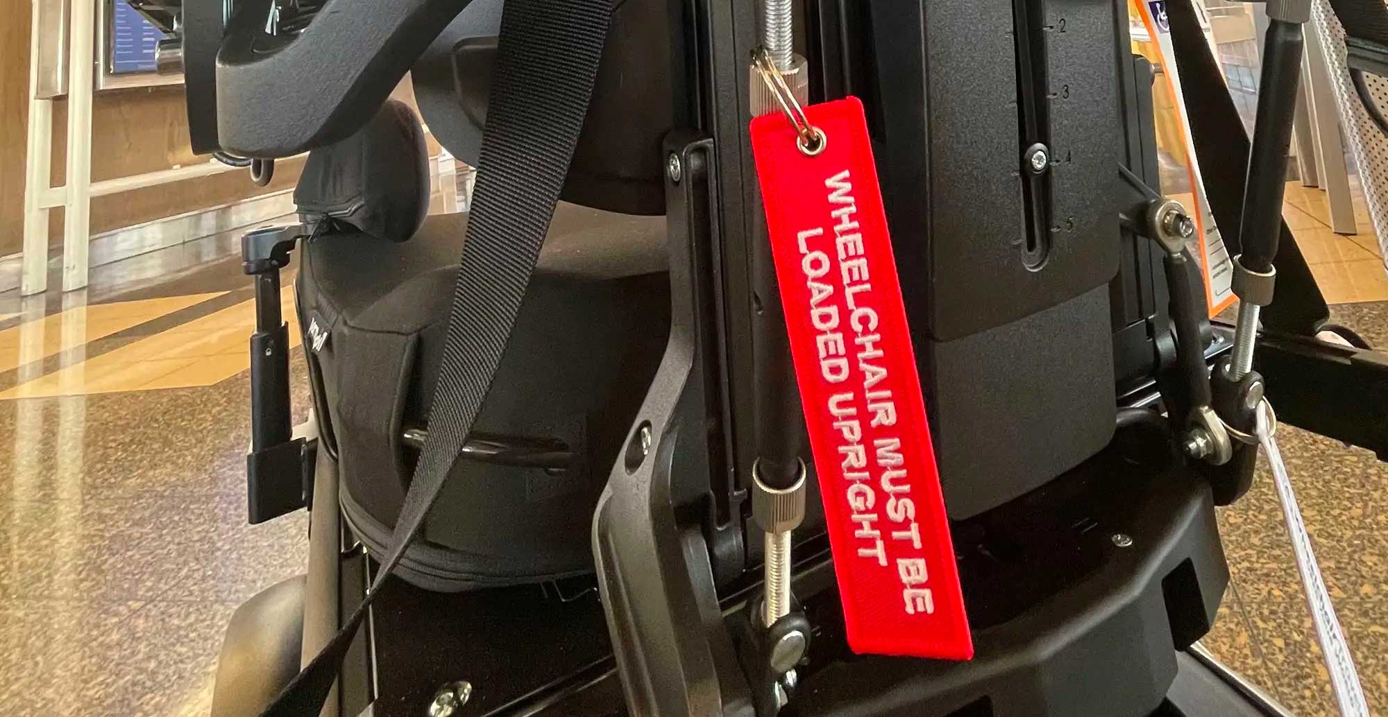 Red fabric luggage tag attached to wheelchair with white embroidered letters that reads wheelchair must be loaded upright.