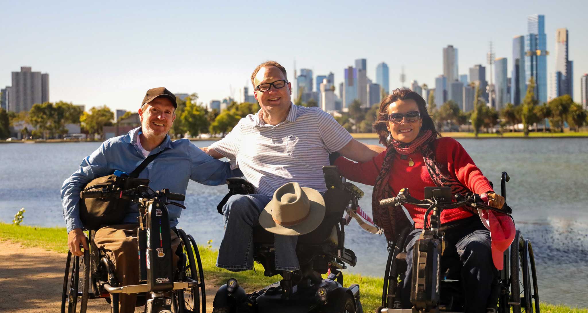 John with two other wheelchair users in front of the Melbourne, Australia skyline.