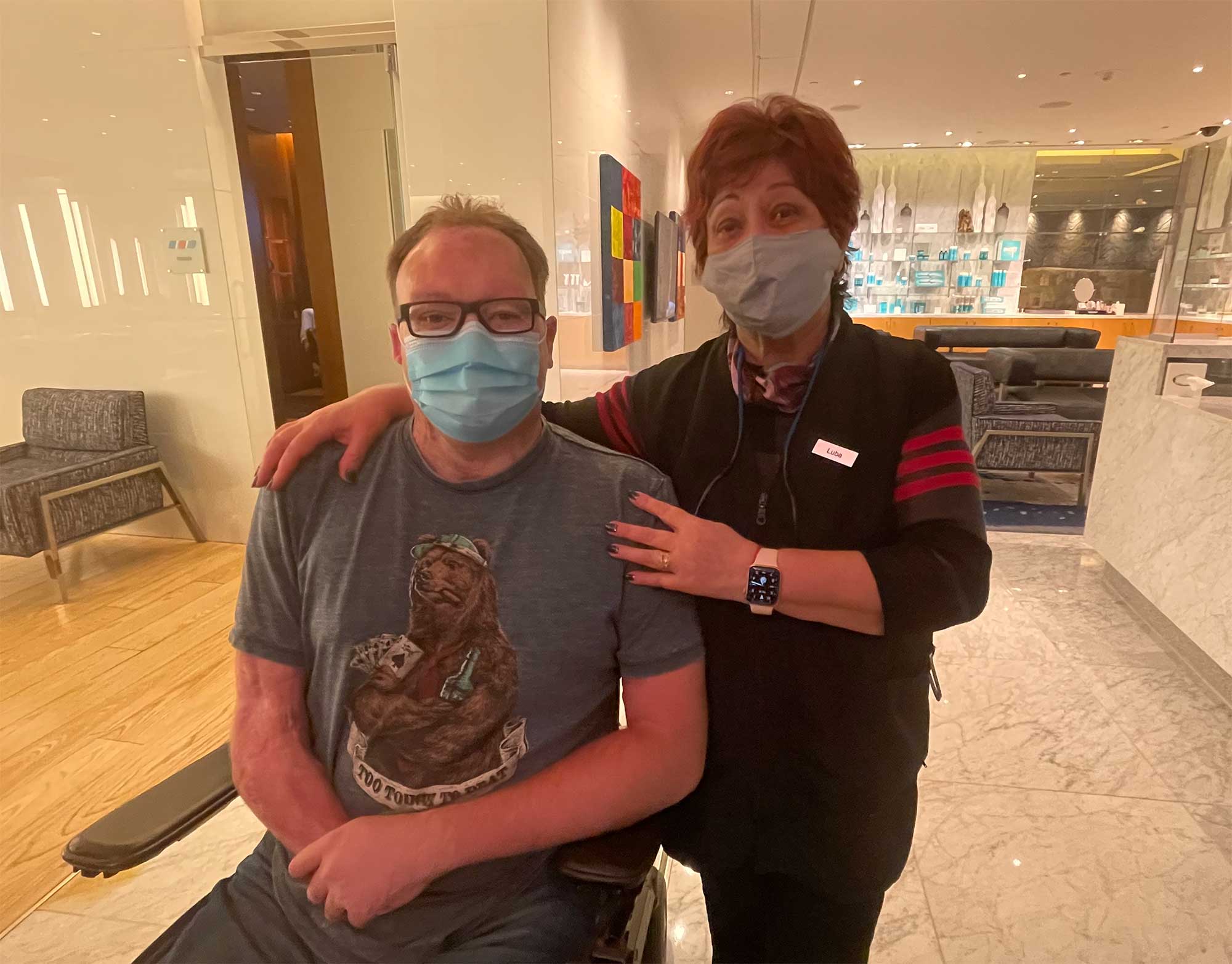 John seated in his wheelchair next to a hair stylist, both are wearing face masks.