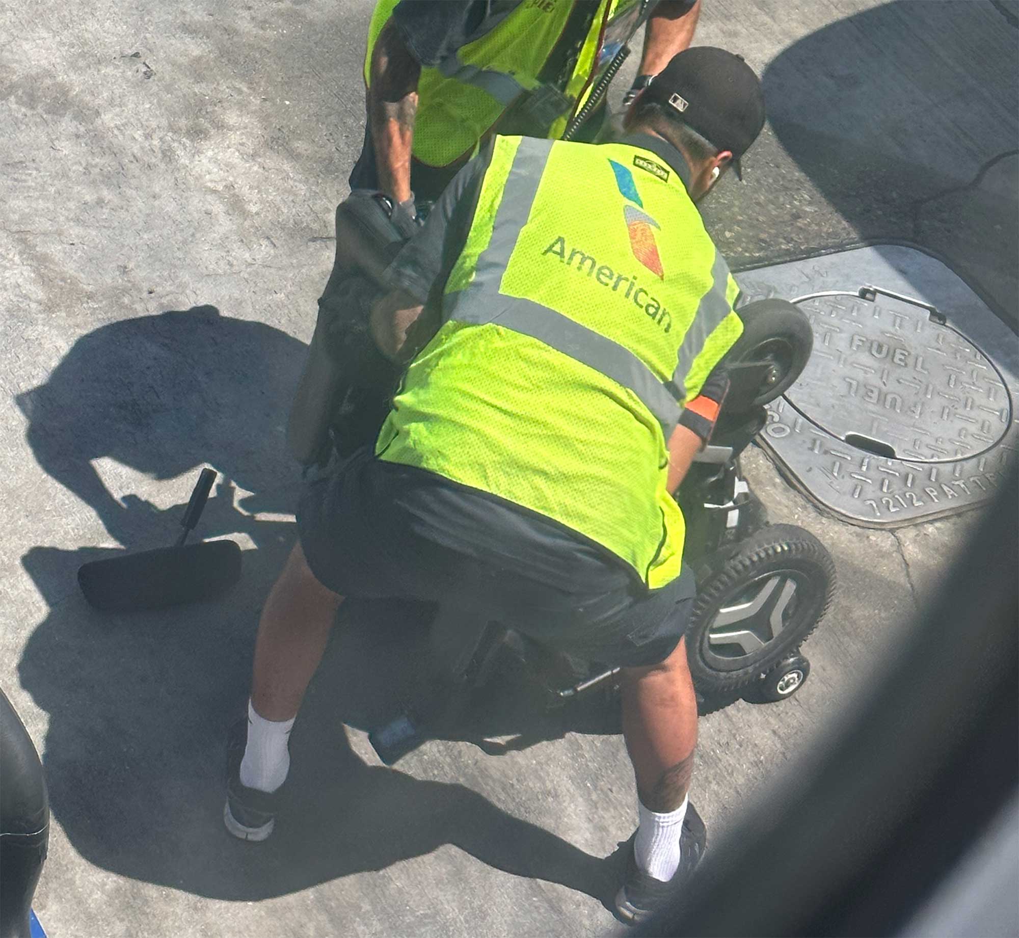 Power wheelchair flipped upside down on the pavement, with two American Airlines ground staff members around it.