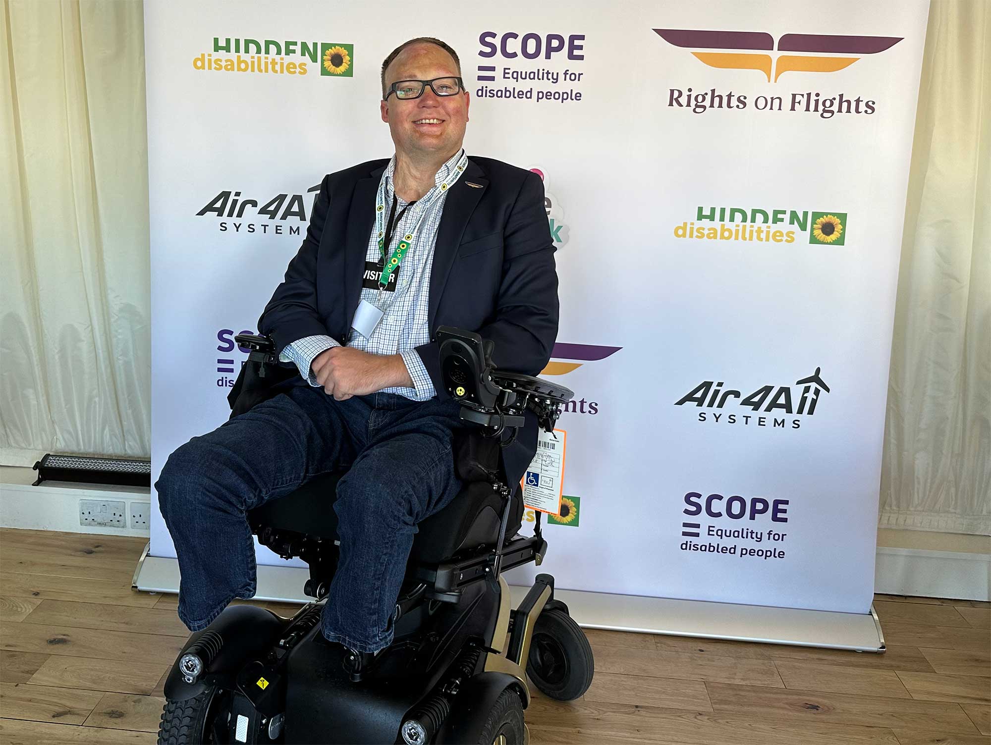 John seated in his wheelchair in front of the Rights on Flight banner that included logos of sponsored including Scope, Air For All, and Hidden Disabilities.