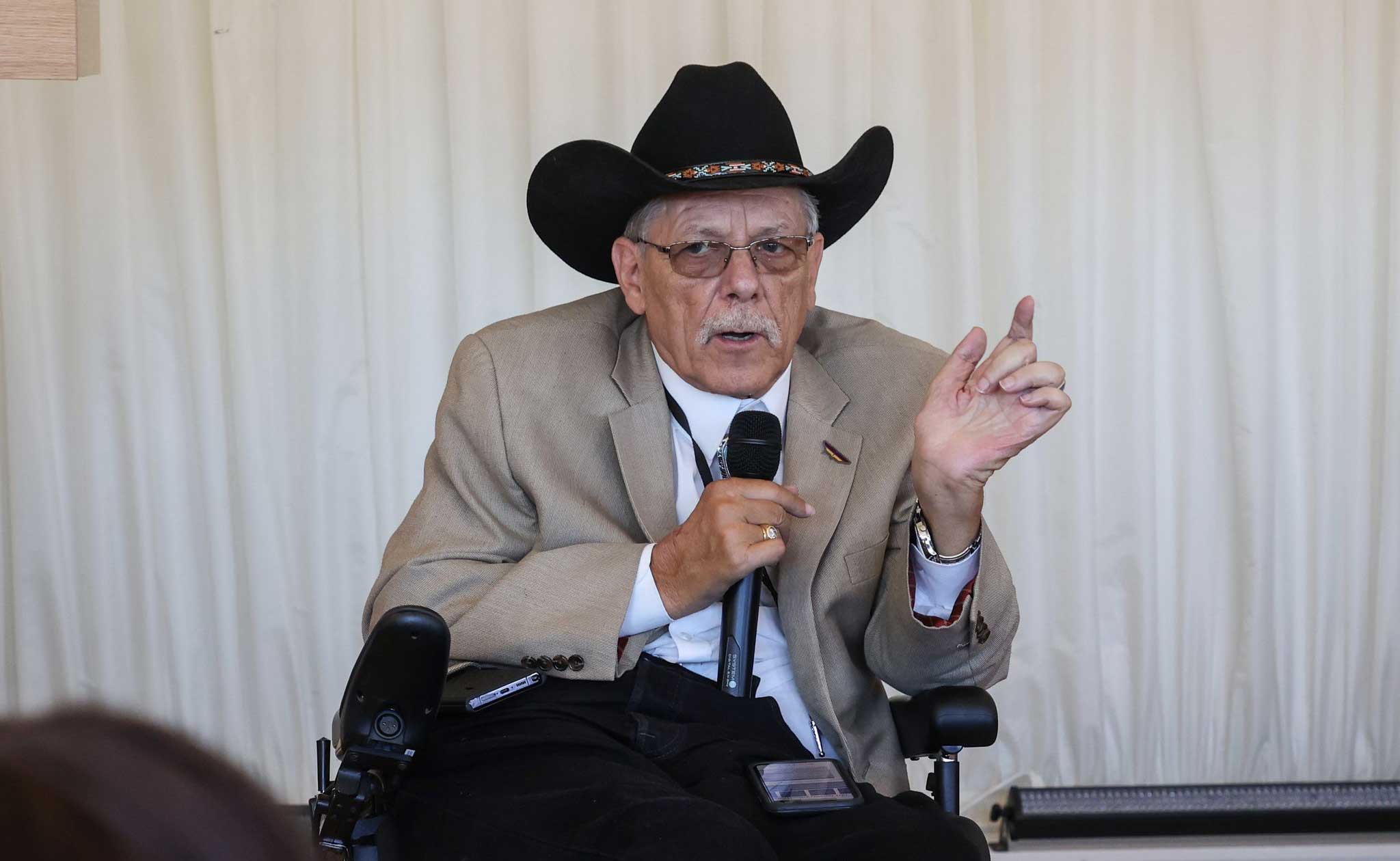 Man seated in wheelchair speaking into a microphone while wearing a cowboy hat.