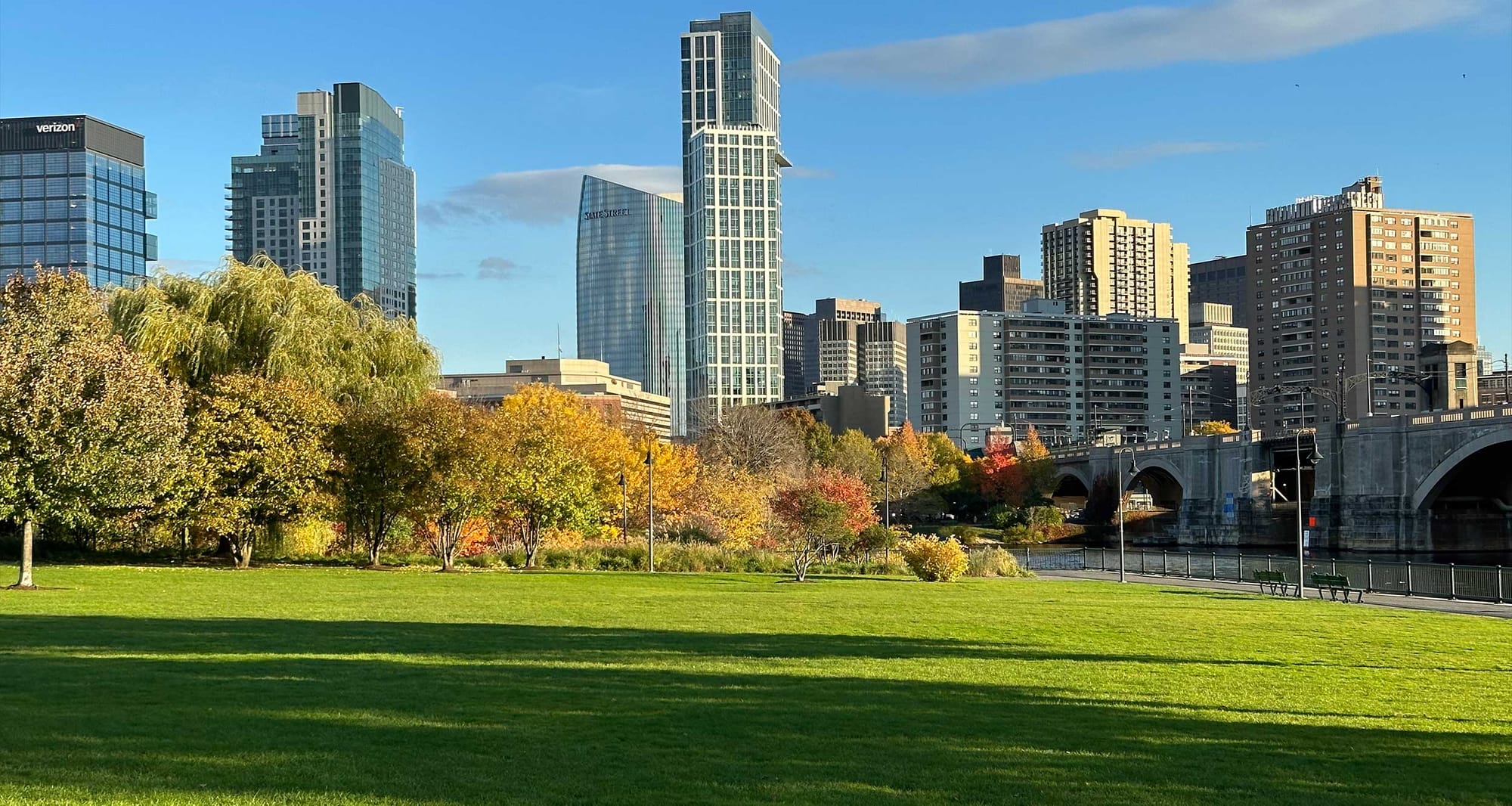 Public park along riverfront, trees with fall foliage and the downtown Boston skyline.