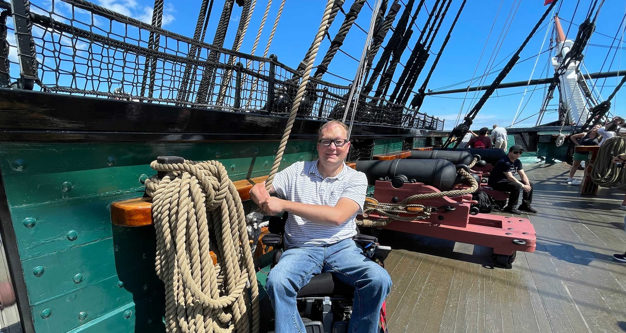 John seated in his wheelchair on the wooden deck of the Constitution with a cannon in the background.