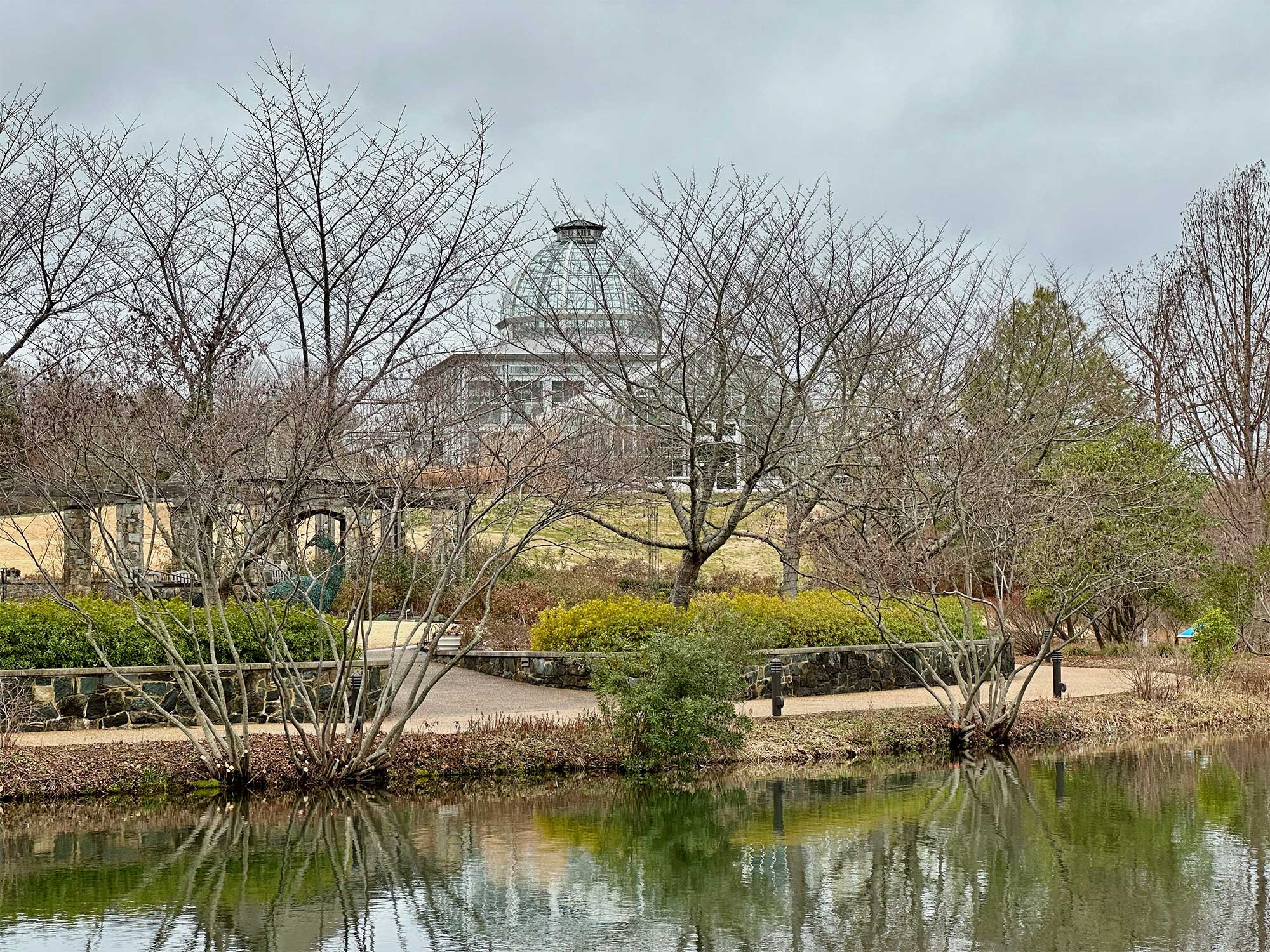 Conservatory building seen through trees with lake in foreground.