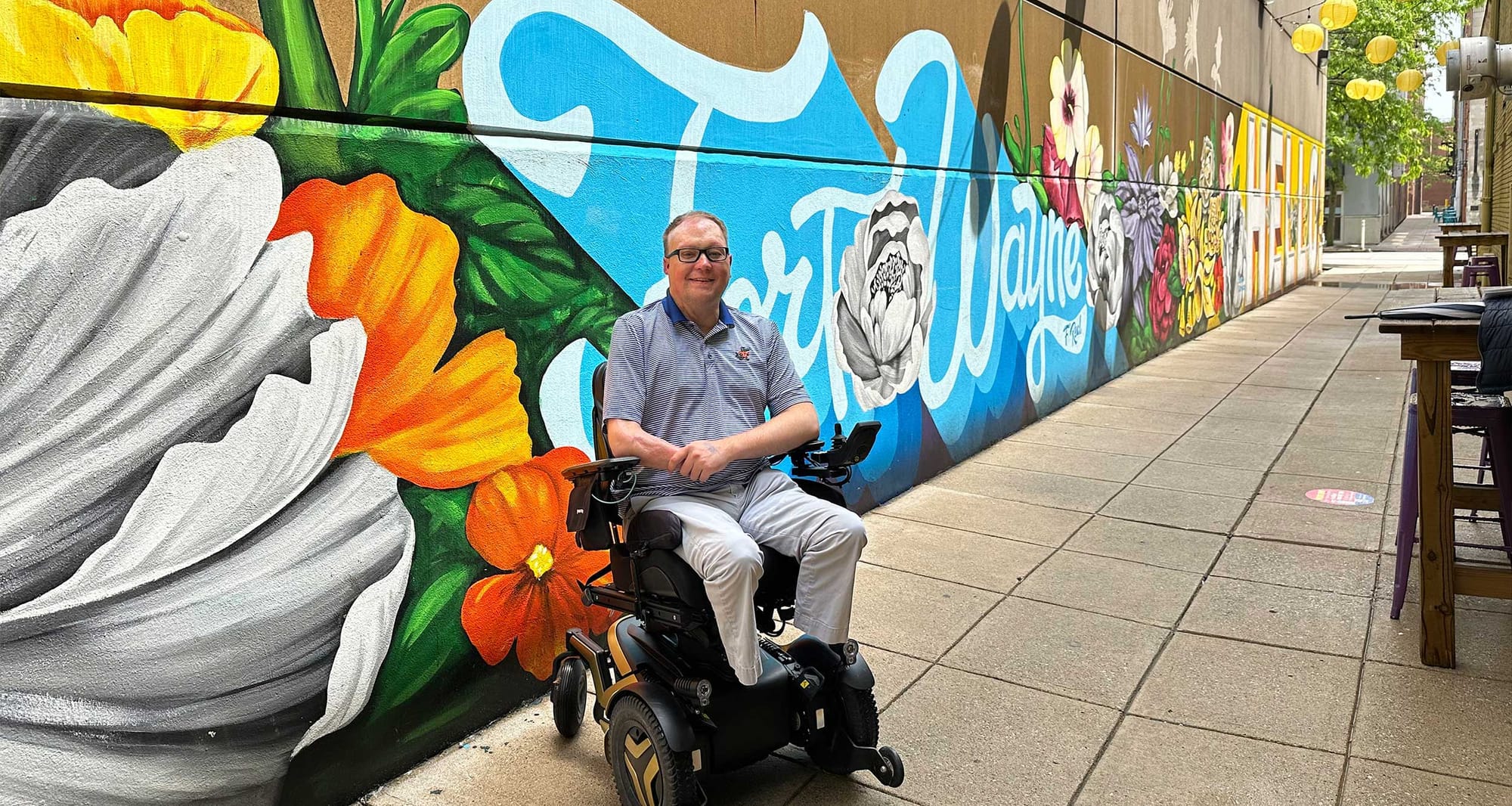 John seated in his wheelchair in an alleyway with a floral mural painted in the words Fort Wayne.