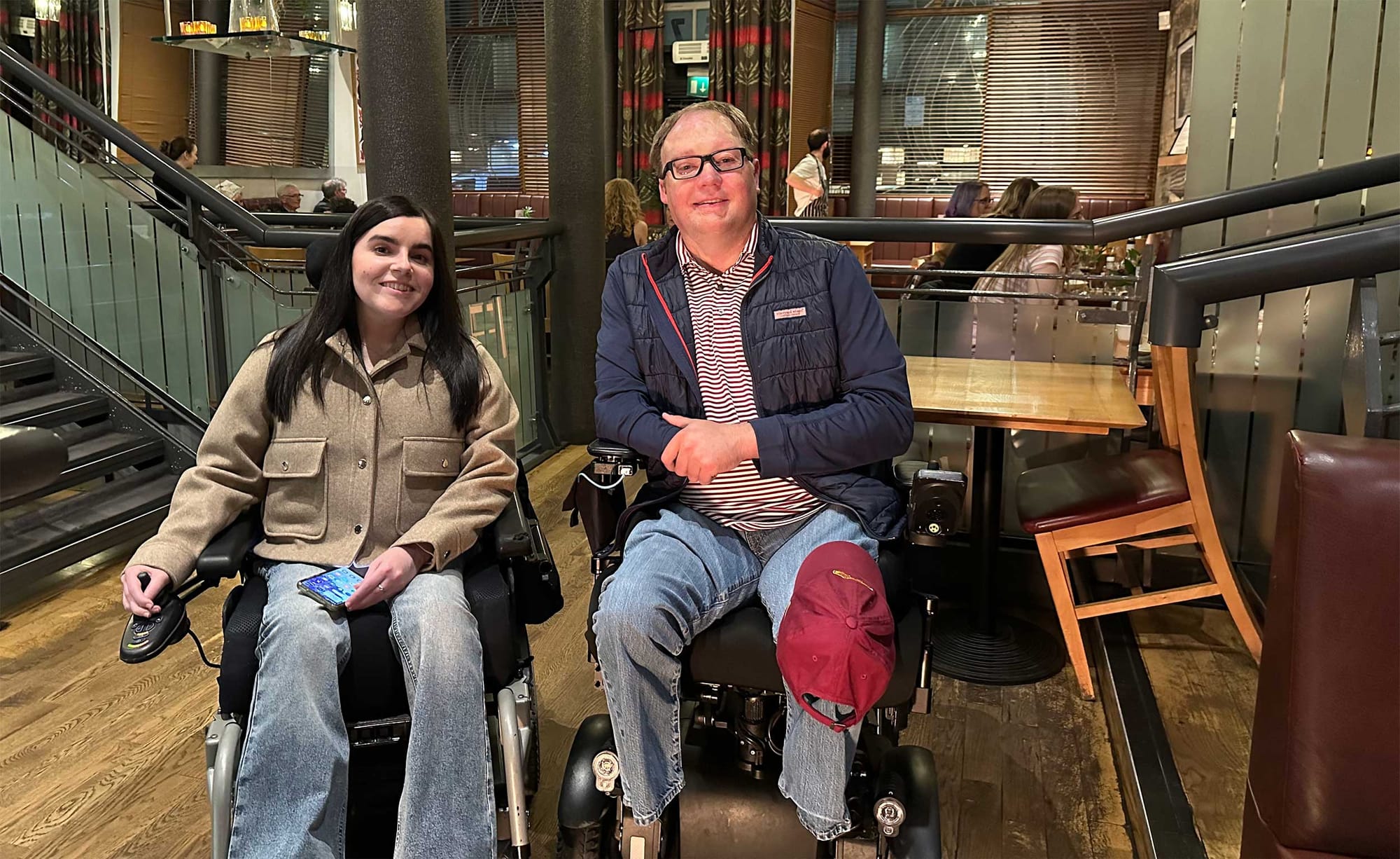 John seated in his wheelchair next to a friend, who also uses a wheelchair, inside a restaurant.