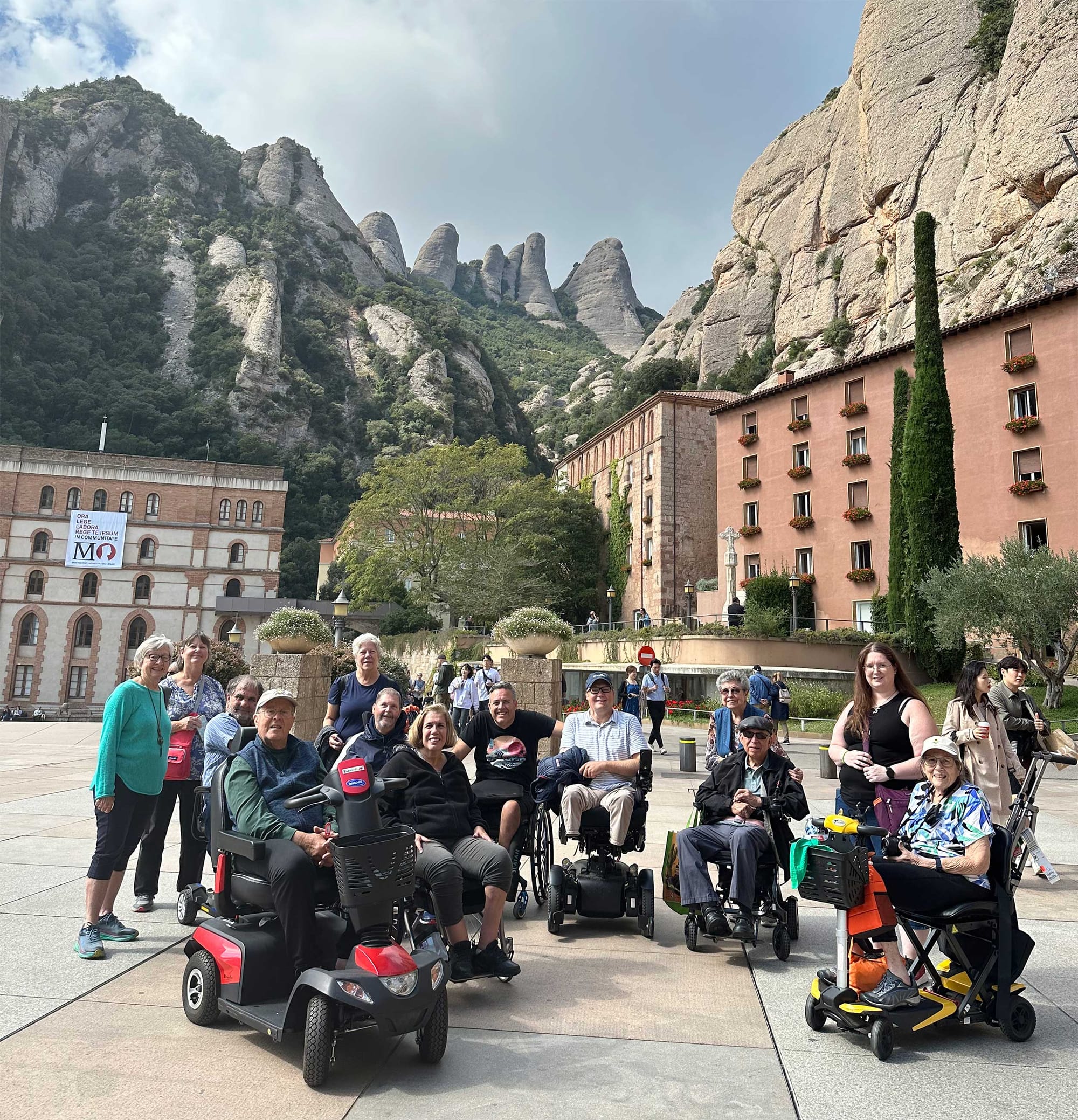 Group of wheelchair and scooter users at foot of large rock formation on mountainside.